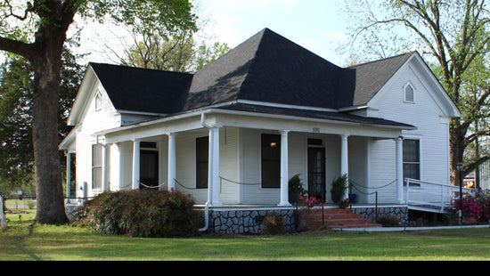 White house with black roof and porch, surrounded by trees and a clear sky